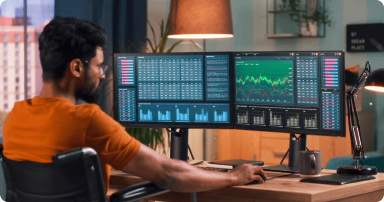 Photo of a man in front of three computer screens displaying various charts and graphs 