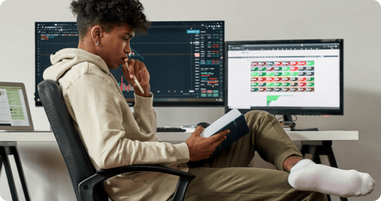 Photo of a man reading a book in front of two computer screens