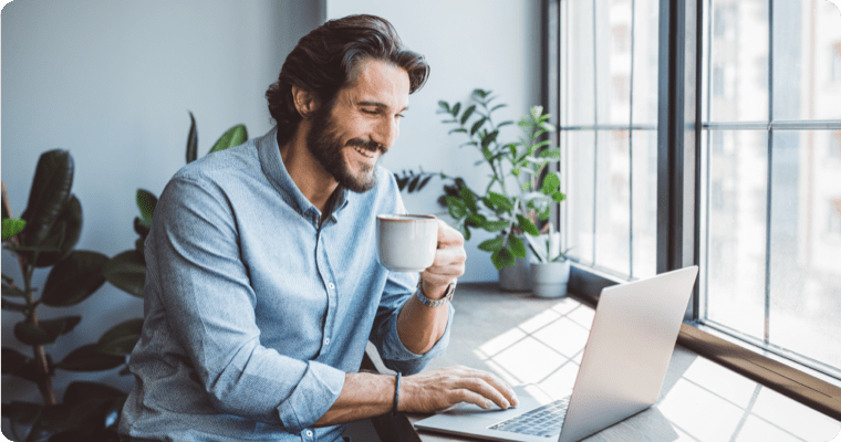 Photo of a smiling man holding a coffee while looking at their computer