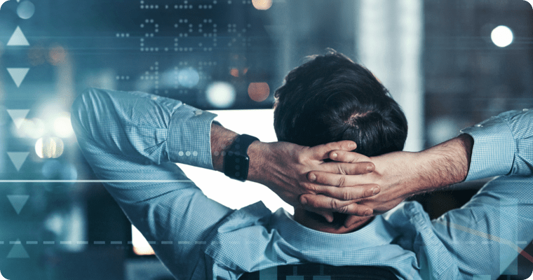 Photo of a man in front of a computer with their arms crossed behind their head