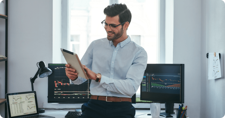 Photo of a smiling man holding a tablet in front of three computer screens
